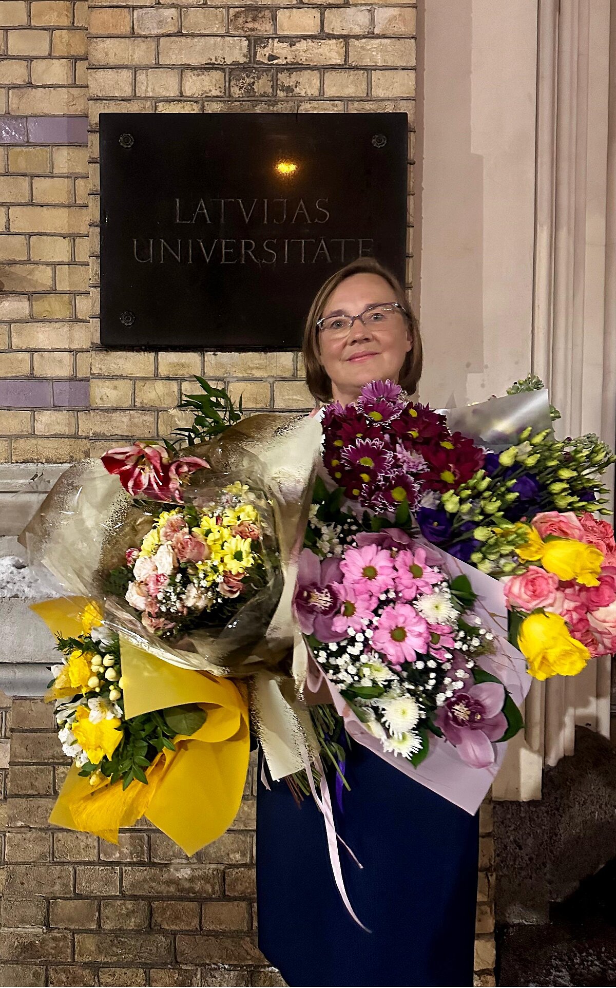 Latvijas Universitātes Madonas filiāles vadītāja Lolita Kostjukova ar filiāles 2026. gada Ziemas absolventu dāvātiem ziediem pie LU ēkas. 23.02.2026. Foto no personīgā arhīva.